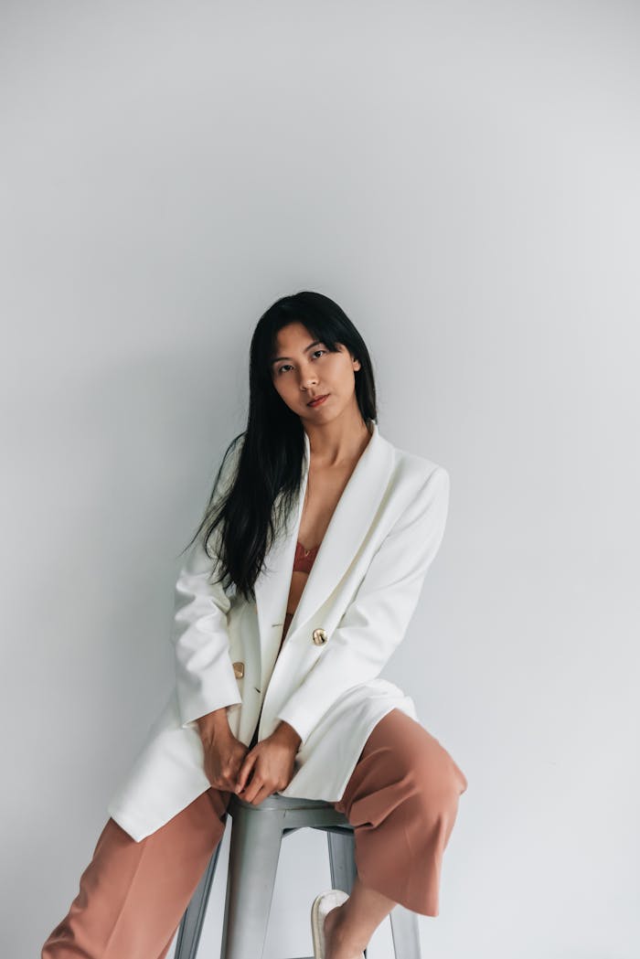 Portrait of an Asian woman in a white blazer sitting on a stool with a minimalistic backdrop.